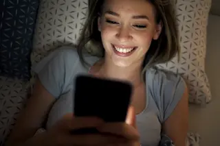 Young woman smiling, lying back in a low lit room with, phone in her hands lighting up her face