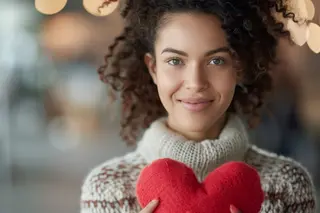 A smiling woman with curly hair holds a pink heart-shaped toy while smiling at the camera in front of a soft blue backdrop