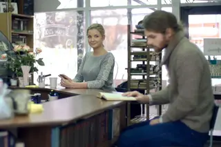 Lady in background smiling while looking at a man reading his book