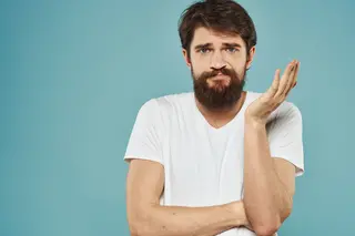 A bearded man with a puzzled expression stands against a blue backdrop