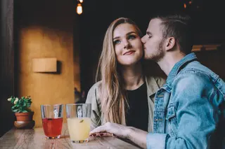 Man kissing smiling woman on the cheek while they sit in a cafe
