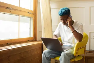 Man sitting beside a window using a phone and with a laptop on his lap