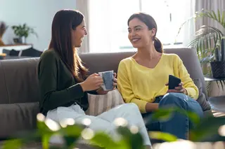 Two smiling young women talking while drinking coffee sitting on the couch at home