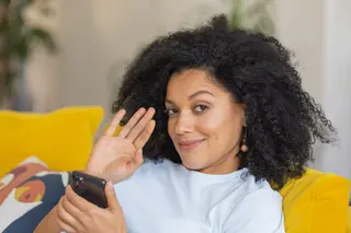 Brunette woman with curly hair sitting on yellow sofa looking at the camera and waving hello