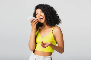 Young woman wearing yellow top in front of grey background recording voice note on smartphone
