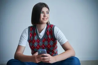 Happy young positive student, phone in hand sitting in front of bright background