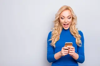 A joyful woman in a stylish blue top discovers exciting news on her smartphone, her face lighting up with surprise and happiness