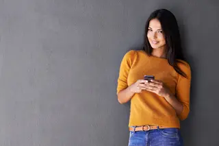 An attractive young woman smiling and holding up a mobile phone while standing against a gray wall